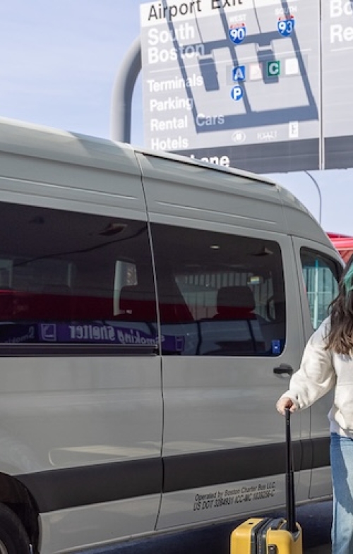 Two women walking into Boston Logan Airport with white shuttle bus in the background