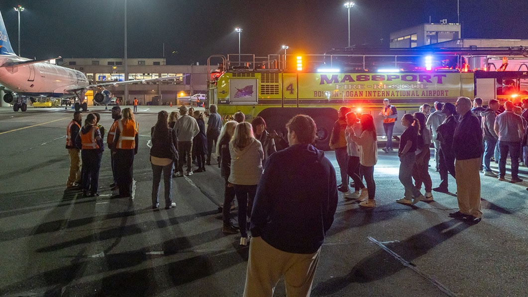 People standing on tarmac in front of Massport fire truck and large plane at nighttime