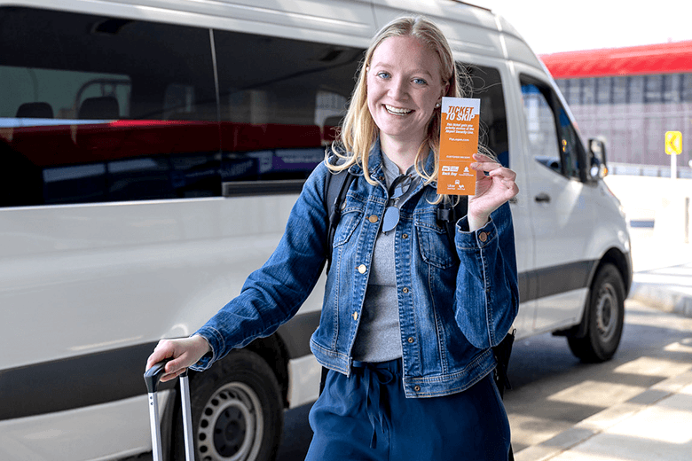 College student holding up ticket to skip flyer standing in front of shuttle bus with the Terminal E red roof in the background